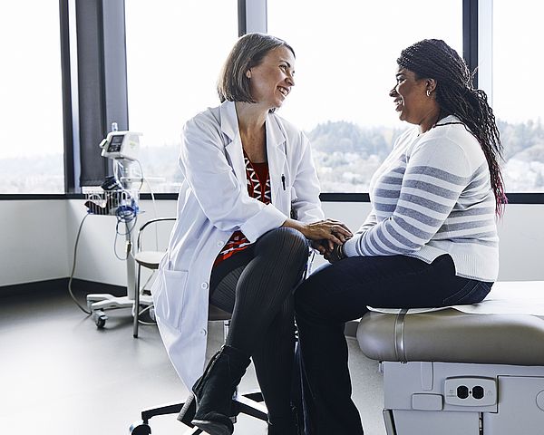 Eine Ärztin und eine Patientin sitzen im Behandlungszimmer gegenüber und unterhalten sich. Die Ärztin hält dabei die Hände der Patientin und spricht ihr gut zu.
