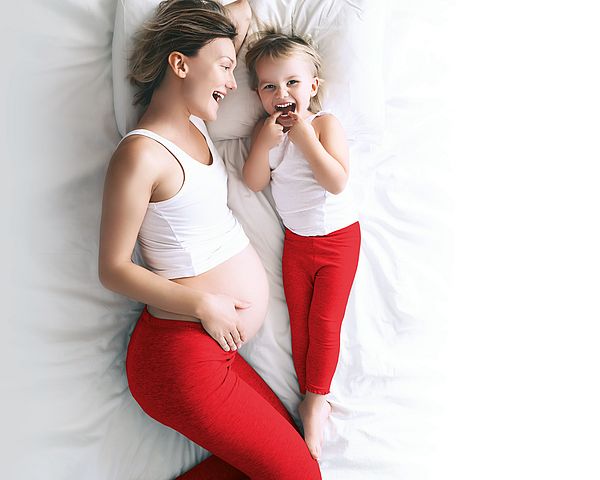 Eine schwangere Mutter liegt mit ihrem Kind lachend im Bett.