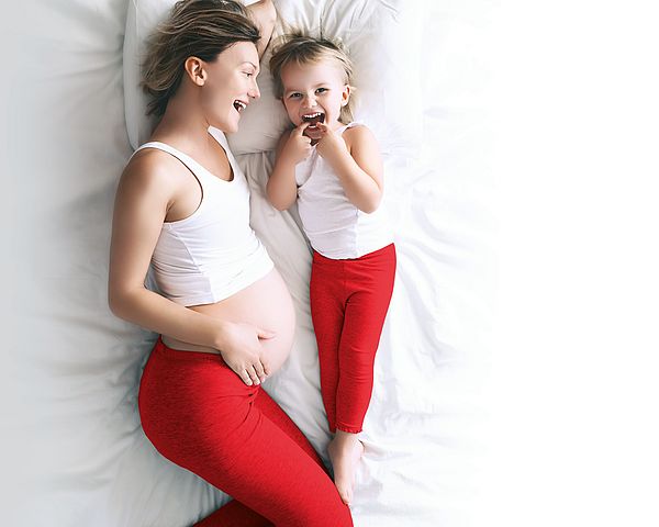 Eine schwangere Mutter liegt mit ihrem Kind lachend im Bett.