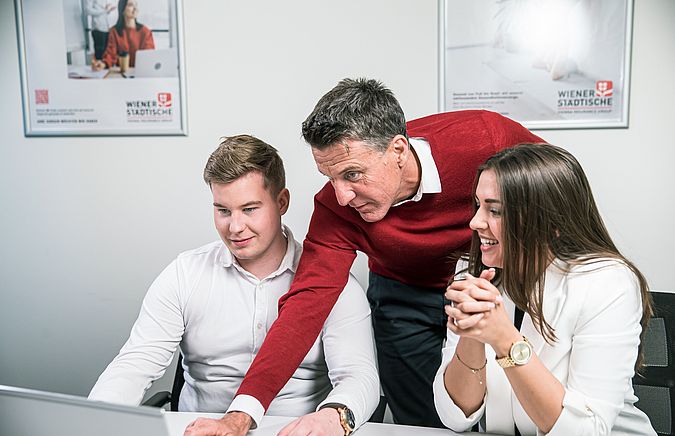 Mann mit rotem Pullover erklärt einem anderen jungen Mann und einer Frau etwas am Laptop.