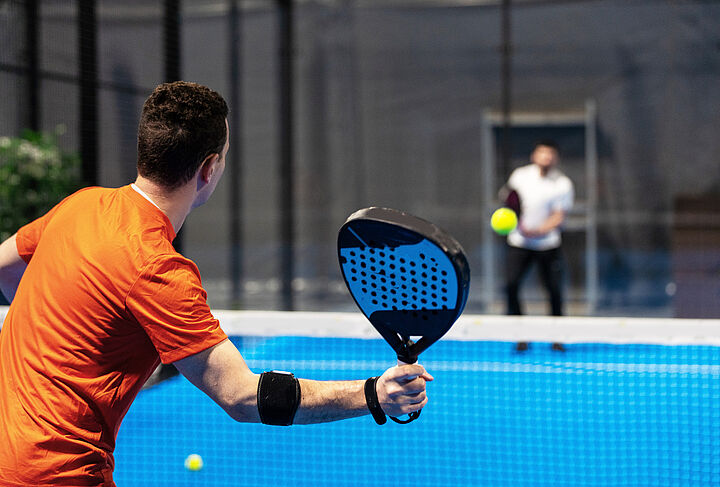 Zwei Männer spielen Padel-Tennis in einem kleinen Court mit leuchtend blauem Boden. Der Mann im Vordergrund trägt ein schwarzes Schweißband am Arm. 