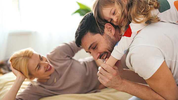 Ein Vater liegt lachend auf dem Bett, während sein kleines Kind ihn umarmt; die Mutter liegt daneben und schaut beide lächelnd an. Die Szene wirkt liebevoll, verspielt und familiär in einer hellen Wohnumgebung.