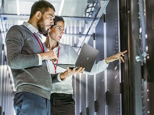 •	Ein Mann und eine Frau der IT-Abteilung stehen mit einem Laptop in einem Rechenzentrum und deuten dabei auf einen Server