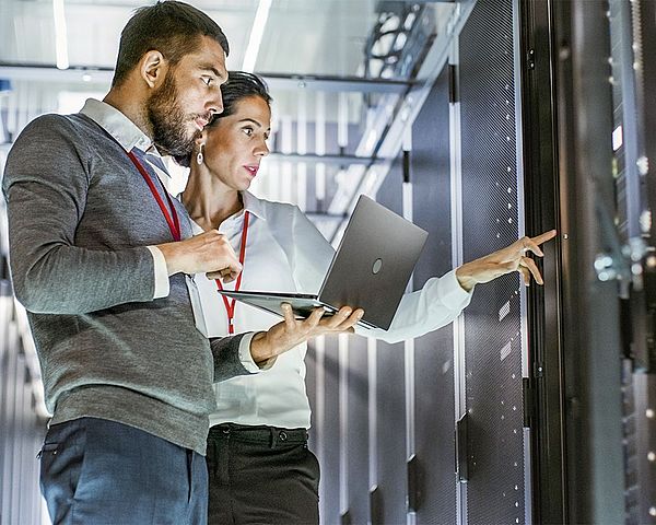 • Ein Mann und eine Frau der IT-Abteilung stehen mit einem Laptop in einem Rechenzentrum und deuten dabei auf einen Server