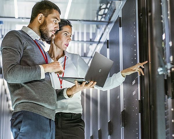 •	Ein Mann und eine Frau der IT-Abteilung stehen mit einem Laptop in einem Rechenzentrum und deuten dabei auf einen Server