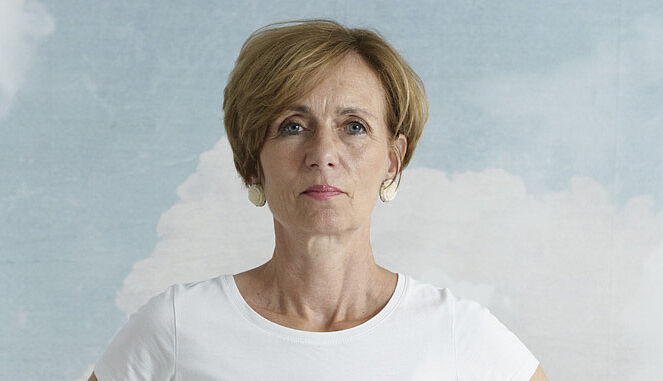 Eine Frau mit kurzen Haaren trägt ein weißes T-Shirt und eine hellgraue Hose. Sie sitzt auf einem Stuhl und zeigt eine Übung vor mit ihren Händen auf dem Bauch.