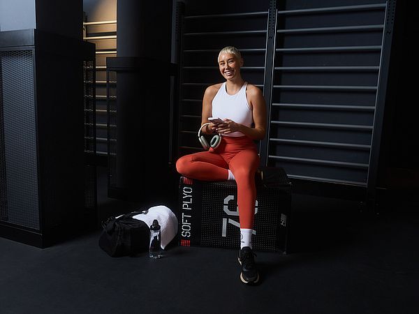 Eine Frau steht im Fitnessstudio. Sie trägt ein weißes Top und rote Leggings, hält ein Handy und eine Trinkflasche, und hat eine schwarze Sporttasche über der Schulter.