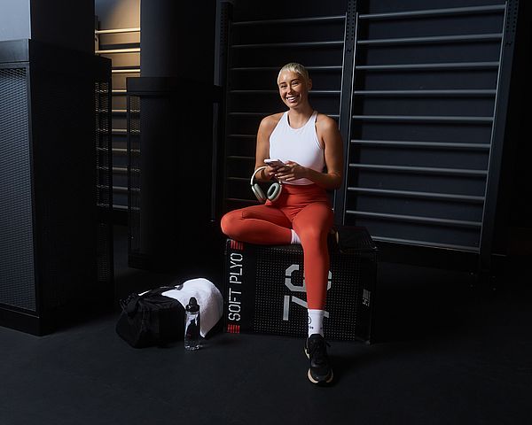 Eine Frau steht im Fitnessstudio. Sie trägt ein weißes Top und rote Leggings, hält ein Handy und eine Trinkflasche, und hat eine schwarze Sporttasche über der Schulter.