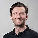 Porträtbild von einem Mann im schwarzen Polo-Shirt mit kurzen dunklen Haaren, Bartstoppeln und einem Lächeln.