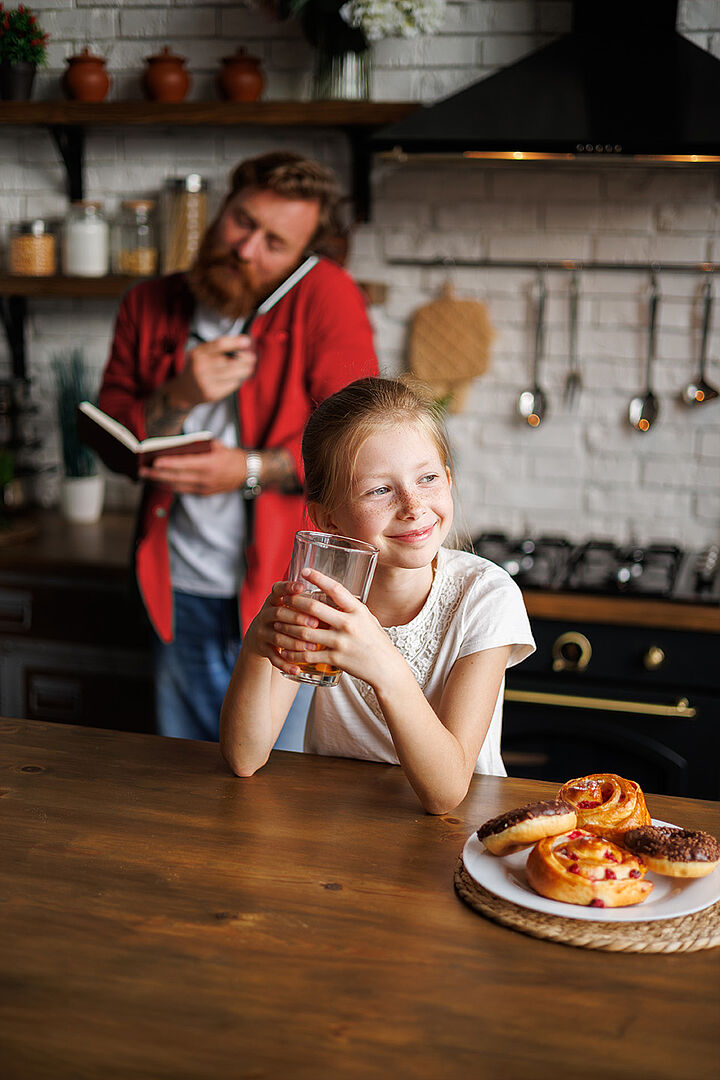 Ein Mädchen sitzt in der Küche am Tisch, hält ein Glas und lächelt. Am Tisch stehen Zimtschnecken und Donuts. Im Hintergrund telefoniert ihr Vater mit einem Notizbuch in der Hand.