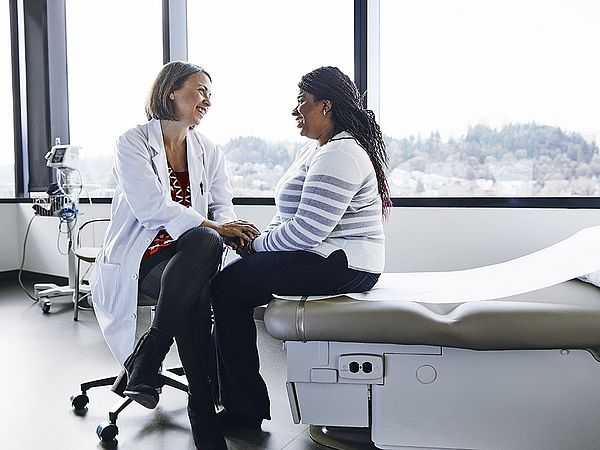 Eine Ärztin und eine Patientin sitzen im Behandlungszimmer gegenüber und unterhalten sich. Die Ärztin hält dabei die Hände der Patientin und spricht ihr gut zu. 