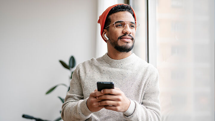 Junger Mann mit Brille, Mütze und Ohrhörern steht am Fenster, hält ein Smartphone und blickt nachdenklich nach draußen.