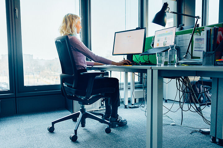 Eine junge Frau mit schwarzer Hose und lila Pullover sitzt auf einem schwarzen Bürosessel an einem Schreibtisch, auf dem ein Bildschirm und ein Laptop aufgebaut sind.