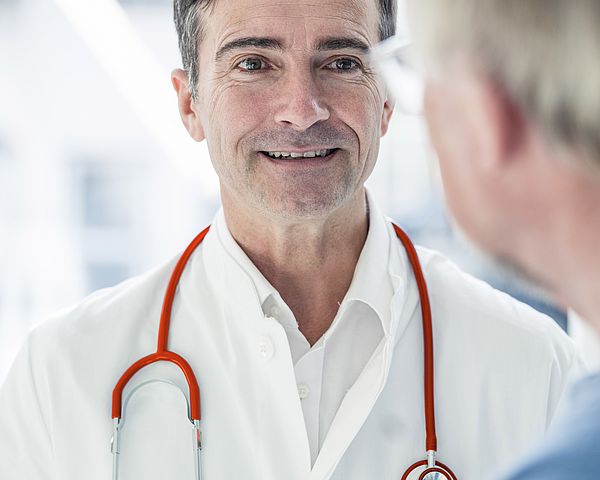 Ein Arzt befindet sich in einer Konversation mit einem älteren Mann und lächelt ihn dabei an. 