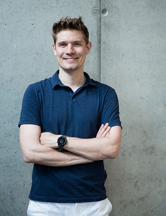 Ein junger Mann steht vor einer Sichtbetonwand, verschränkt die Arme und lächelt. Er hat kurzes braunes Haar, er trägt ein dunkelblaues Poloshirt und eine weiße Hose.