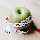 Ein grüner Apfel liegt auf einer Holztischplatte und ist von einem weiß-roten Maßband umwickelt.