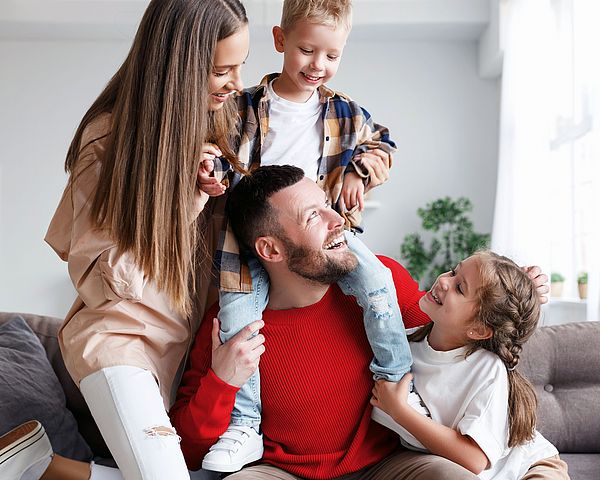 Eine junge glückliche Familie sitzt zu Hause auf der Couch. Der Sohn sitzt auf den Schultern seines Vaters und die beiden Töchter sitzen neben ihm