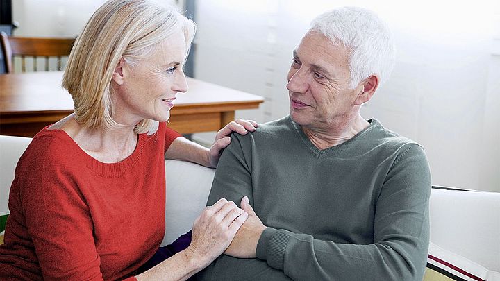 Ein älteres Paar sitzt nebeneinander auf der Couch. Die Frau hält die Hand ihres Mannes.