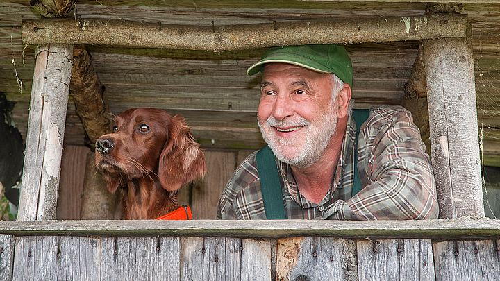 Ein Jäger ist mit seinem Hund am Hochstand im Wald und blickt in die Ferne.