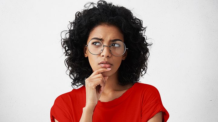 Eine Frau, mit lockigem Haar und Brille, fasst sich an das Kinn und setzt einen nachdenklichen Blick auf