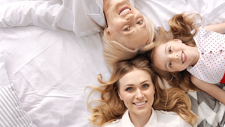 Drei Frauen verschiedener Generationen liegen Kopf an Kopf lächelnd im Bett.
