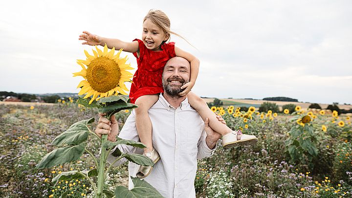 Ein Vater trägt seine junge Tochter lachend auf den Schultern. Dabei spazieren sie durch ein Blumenfeld und beide greifen nach einer Sonnenblume.   