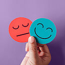 Eine Hand hält zwei kleine, kreisrunde Papierschilder: das blaue zeigt ein Smiley, das rote ein Emoji mit geradem Mund. 