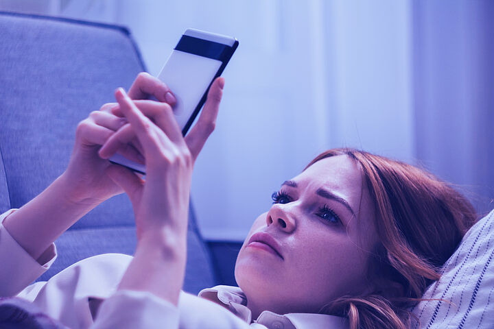 Eine junge Frau mit braunen Haaren hält im Liegen ein Smartphone in der Hand und schaut bekümmert drein.