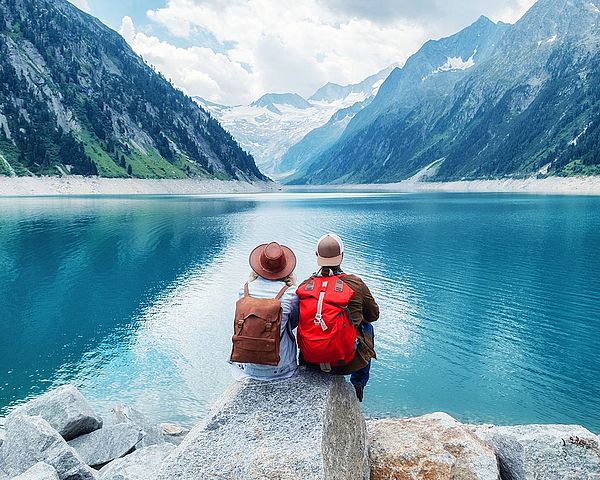 Reisendes Paar sitzt auf einem Felsen und blickt auf einen See in den Bergen.