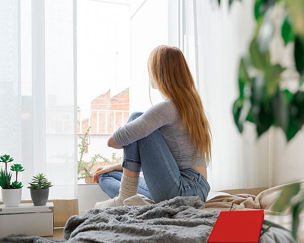 Eine junge Frau sitzt auf ihrem Bett und schaut aus dem Fenster.