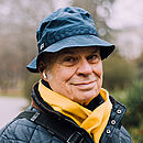 Porträtfoto von einem älteren Mann mit dunkler Winterjacke, gelbem Schal und blauem Fischerhut, im Hintergrund der Gehweg in einem Park und mehrere Bänke.