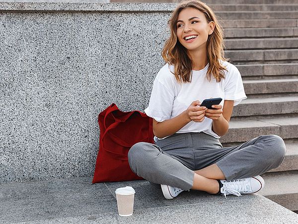 Eine junge Frau sitzt auf Treppen im Freien und tippt auf ihrem Smartphone.