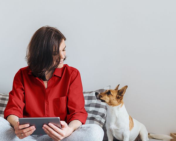 Eine junge Frau sitzt lächelnd mit ihrem Tablet und ihrem Hund im Bett.