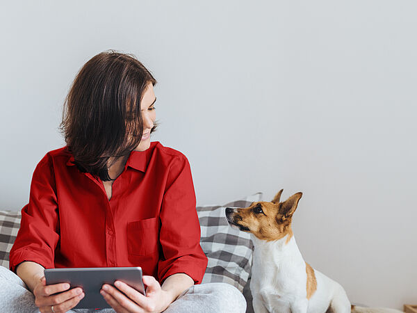 Eine junge Frau sitzt lächelnd mit ihrem Tablet und ihrem Hund im Bett.