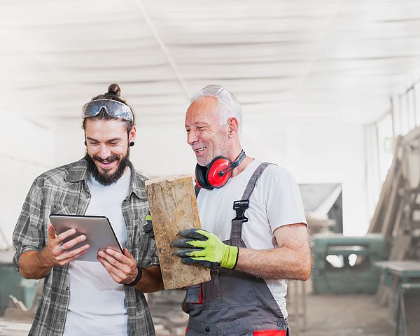 Ein älterer Mann mit einem Stück Holz in der Hand und ein junger Mann blicken auf ein Tablet einer Werkstatt.
