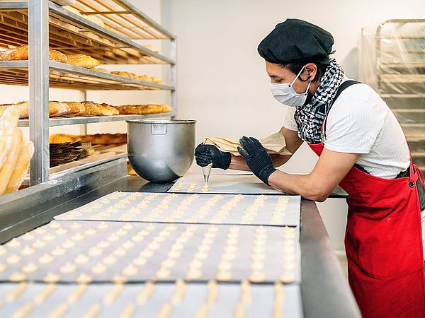 •	Ein Konditormeister steht in seiner Backstube mit Maske und Handschuhen. Dabei formt er voller Konzentration diverse Gepäcksorten. 