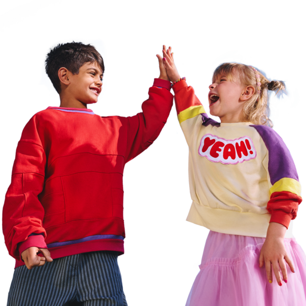 Zwei lachende Kinder geben sich ein High Five. Ein Kind trägt einen bunten Pullover mit der Aufschrift ‚YEAH!‘, das andere einen roten Pullover.