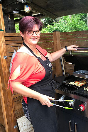 Sandra Schöckenfuchs, eine Außendienstmitarbeiterin der Wiener Städtischen, steht hinter einem Grill in ihrem Garten und bereitet gerade Gemüse zu. Dabei trägt sie eine Schürze und lächelt in die Kamera