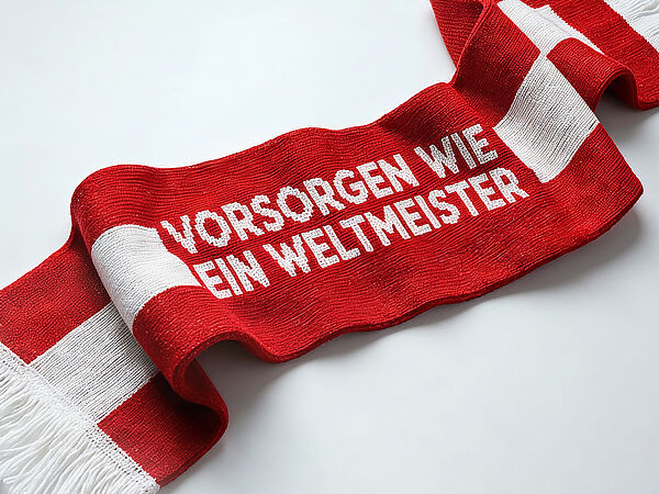 Roter Schal mit weißen Streifen liegt locker gewellt auf hellem Hintergrund. Auf dem Schal steht in großen weißen Buchstaben „Vorsorgen wie ein Weltmeister“.
