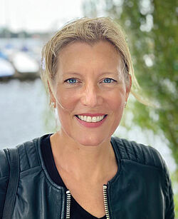 Porträtbild einer Frau mit blonden, zusammengebundenen Haaren in schwarzer Lederjacke, dahinter viel Grün und eine Wasseroberfläche.