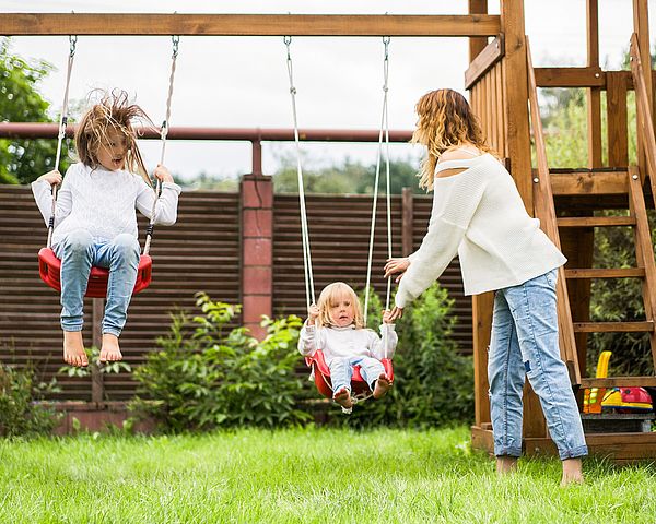 Zwei Kinder sitzen auf einer Schaukel und werden von einer jungen Frau angestoßen.