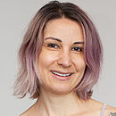Eine junge Frau sitzt auf einem grauen Behandlungsstuhl, lächelt und hat auffällige Tattoos an den Armen.
