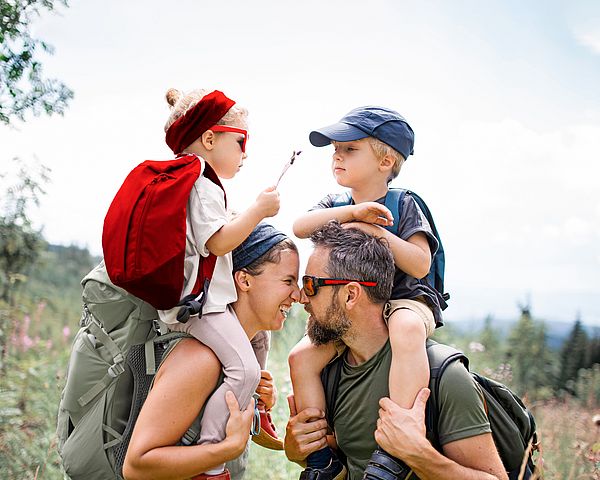 Eine junge glückliche Familie mit zwei Kindern wandert in den Bergen. Die Kinder werden auf den Schultern getragen. 