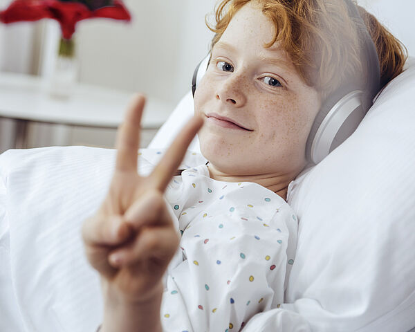 Ein Kind liegt im Krankenbett mit Kopfhörer und zeigt dabei lächelnd ein Peace-Zeichen