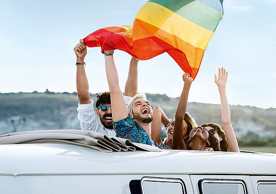 Junge Menschen schauen beim Schiebedach von Auto raus und schwingen eine Regenbogenfahne.