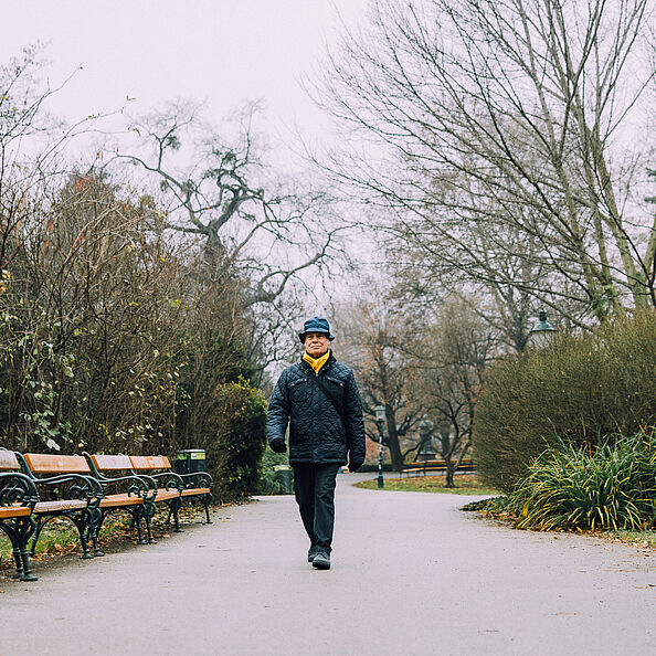 Ein Mann spaziert am Gehweg im Park, vorbei an Parkbänken, vor herbstlicher Vegetation.