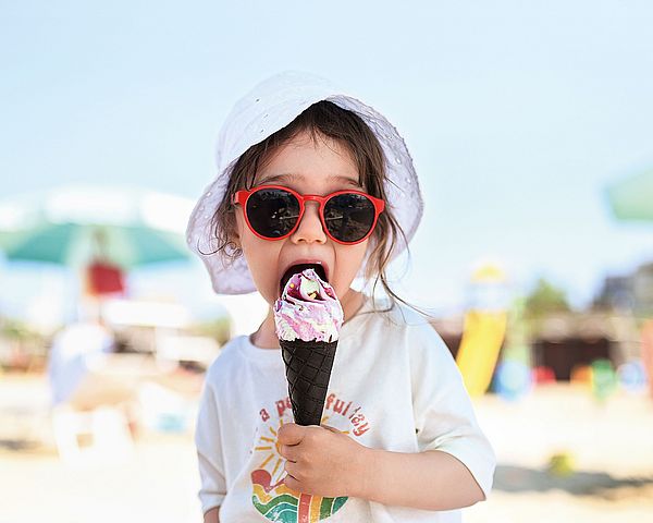 Ein junges Mädchen mit Sonnenhut und Brille isst am Strand ein Eis.