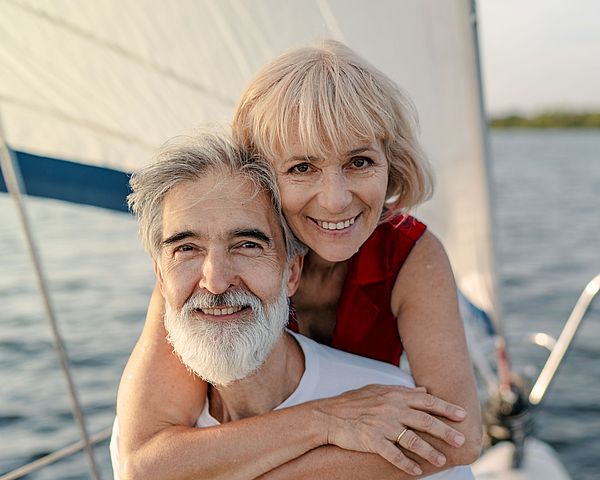 Ein glückliches Seniorenpaar steht auf einem Segelschiff am Meer. Die Frau umarmt den Mann von hinten.