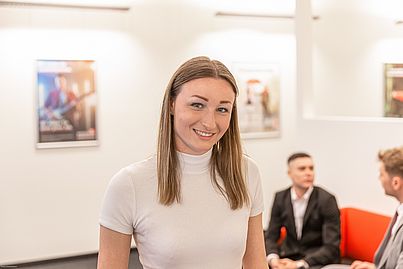 Junge Frau mit weißem kurzärmeligen Rollkragenpullover lächelt. Im Hintergrund sieht man zwei Männer die sich unterhalten.