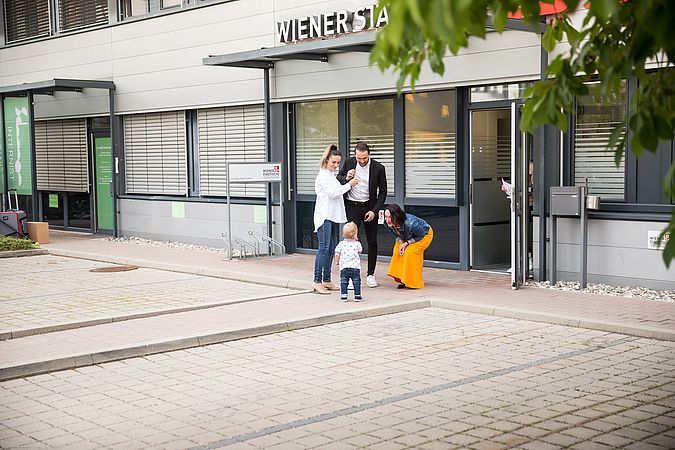 Ein Mann und zwei Frauen blicken zu einem kleinen Kind. Die Personen befinden sich vor einer Wiener Städtischen Geschäftsstelle.